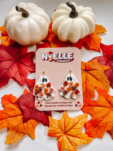 Pair of ghost-shaped earrings on a 'Noelle Earrings' card with fall leaves in the background